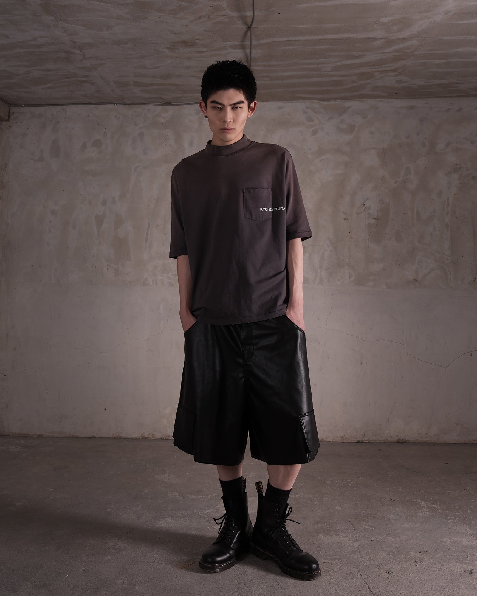 KYOHEY_FUJITAのヘビーウォッシュドモックネックロゴTシャツを着用した男性モデル。ダークグレーの半袖Tシャツに黒のレザー風ハーフパンツを合わせたストリートスタイル。